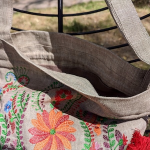 May include: A beige linen tote bag with a red tassel and embroidered floral design featuring red, orange, green, and purple flowers.