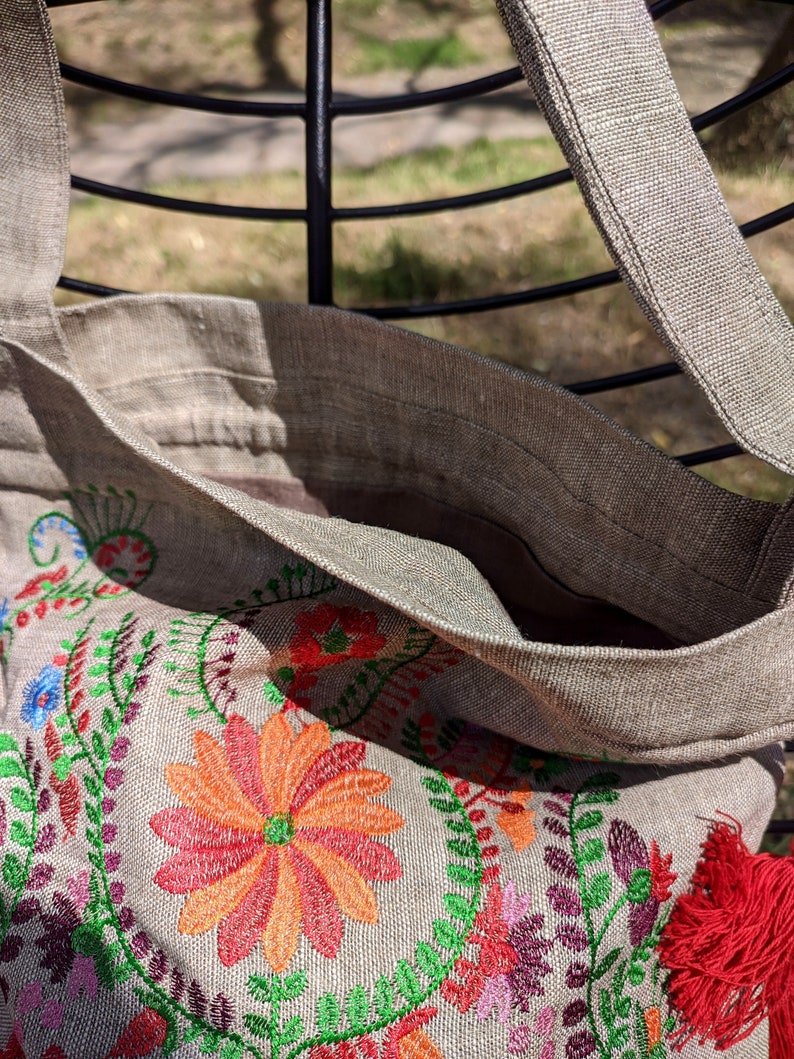 May include: A beige linen tote bag with a red tassel and embroidered floral design featuring red, orange, green, and purple flowers.