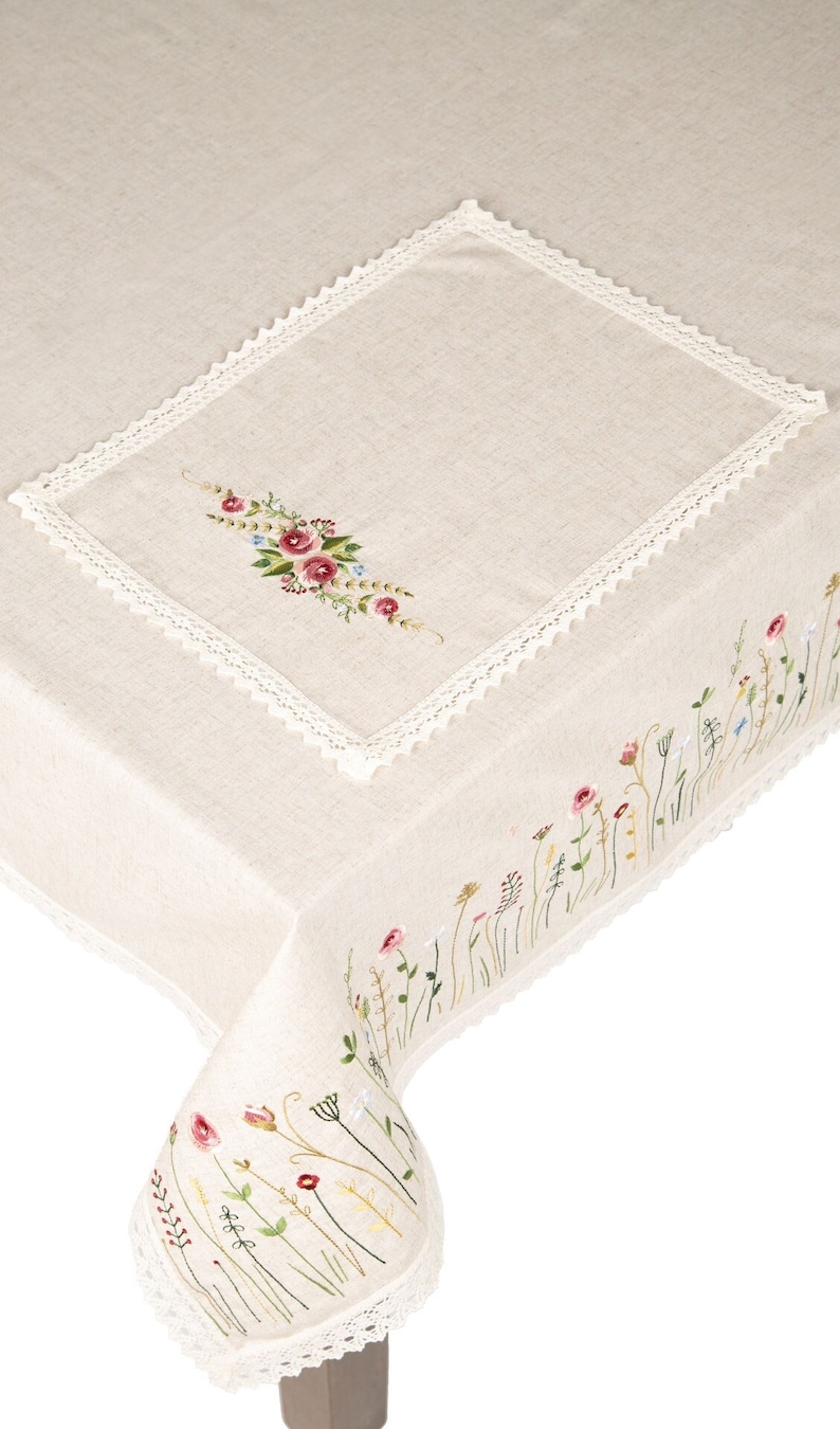 May include: A beige linen tablecloth with a white crocheted border and embroidered floral designs. The tablecloth features a bouquet of flowers in the center and a row of smaller flowers along the edge.