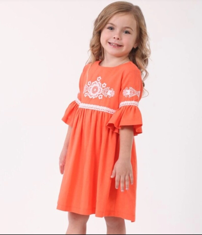 K&ouml;nnte beinhalten: Ein junges M&auml;dchen tr&auml;gt ein leuchtend oranges Kleid mit wei&szlig;en gestickten Blumen und einer wei&szlig;en Spitzenborte. Das Kleid hat einen leicht ausgestellten Rock und kurze &Auml;rmel.