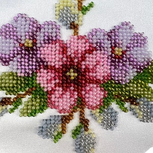 K&ouml;nnte beinhalten: Nahaufnahme eines Perlen-Blumenmusters auf einem wei&szlig;en Stoff. Das Muster besteht aus rosa, lila und gr&uuml;nen Perlen, die einen Blumenstrau&szlig; mit Bl&auml;ttern bilden.