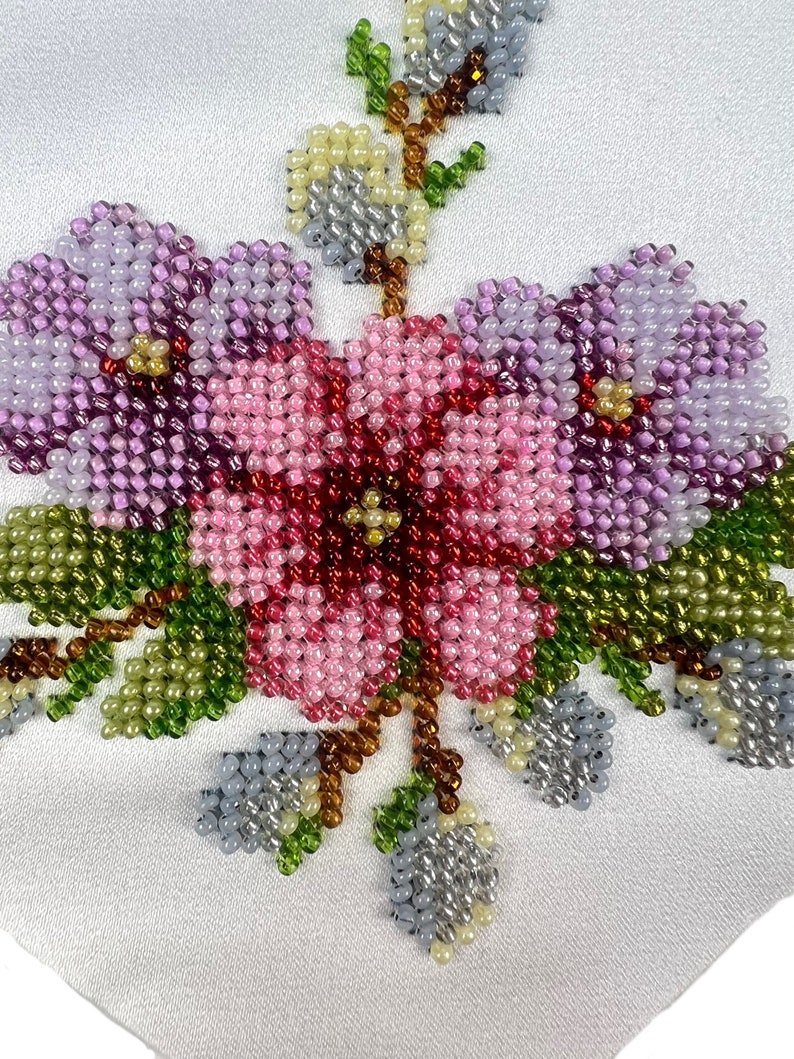 K&ouml;nnte beinhalten: Nahaufnahme eines Perlen-Blumenmusters auf einem wei&szlig;en Stoff. Das Muster besteht aus rosa, lila und gr&uuml;nen Perlen, die einen Blumenstrau&szlig; mit Bl&auml;ttern bilden.