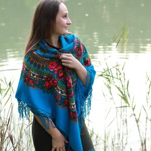 May include: A vibrant blue shawl with a floral pattern featuring red roses and green leaves. The shawl has a fringed edge and is draped over the shoulders. The background shows a body of water and tall grass.