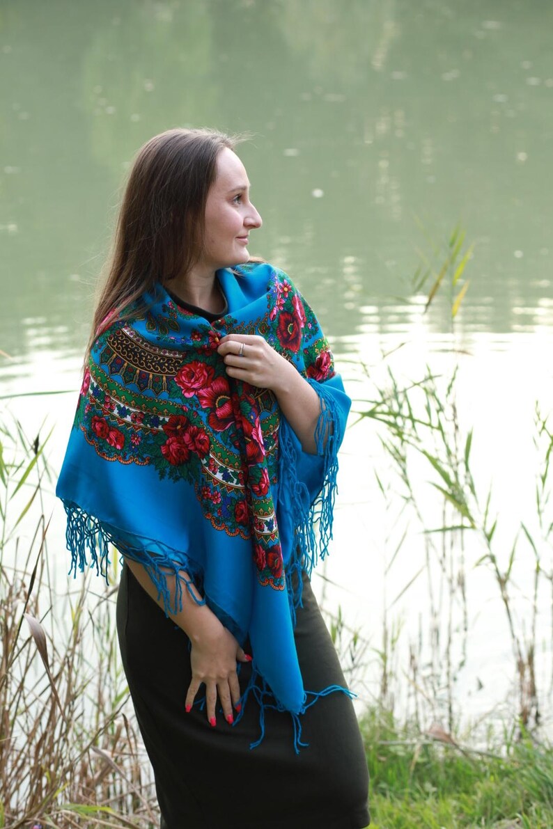 May include: A vibrant blue shawl with a floral pattern featuring red roses and green leaves. The shawl has a fringed edge and is draped over the shoulders. The background shows a body of water and tall grass.