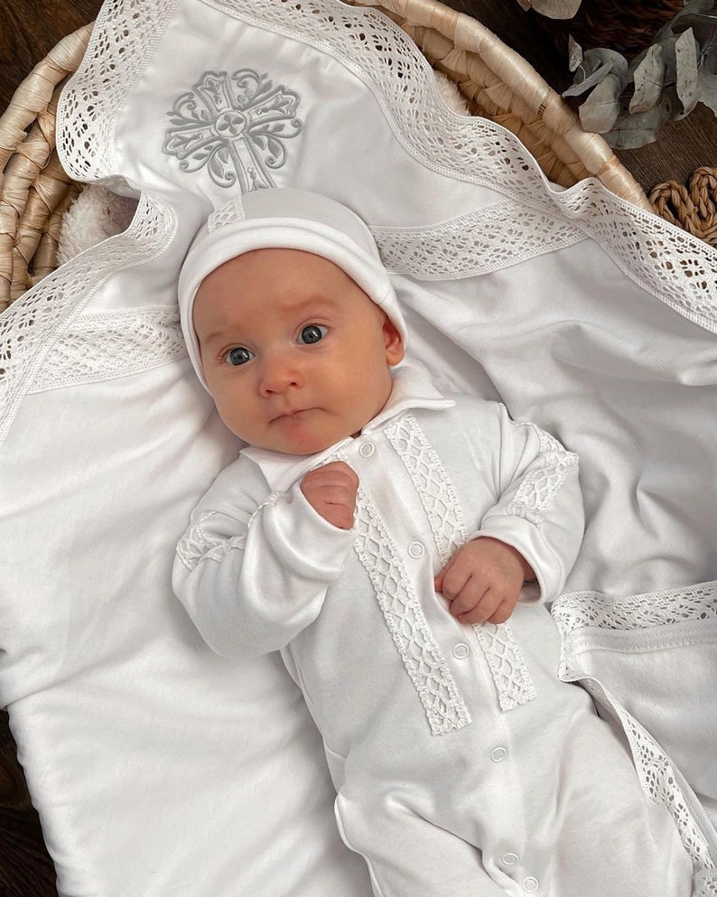 May include: A white baby onesie with lace trim and a white crocheted hat. The onesie has a cross embroidered on the front. The baby is lying in a wicker basket with a white blanket.