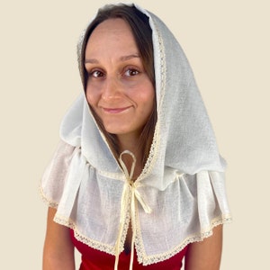 May include: A white linen hood with a cream-colored lace trim and a matching tie closure. The hood is worn over the shoulders and frames the face. The garment is a historical or costume piece.