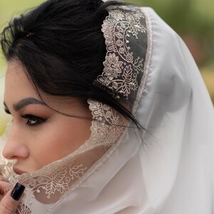 May include: A white veil with intricate floral lace trim. The veil is draped over the head and shoulders, with the lace detail visible along the edge. The background is blurred, suggesting an outdoor setting.