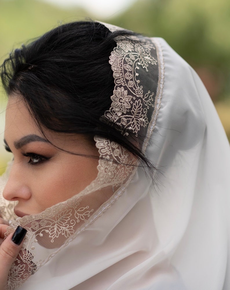 May include: A white veil with intricate floral lace trim. The veil is draped over the head and shoulders, with the lace detail visible along the edge. The background is blurred, suggesting an outdoor setting.