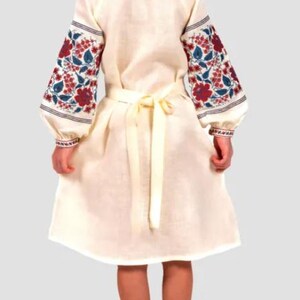 May include: A white linen dress with embroidered floral patterns in red, blue, and green on the sleeves. The dress has a tie belt at the waist.