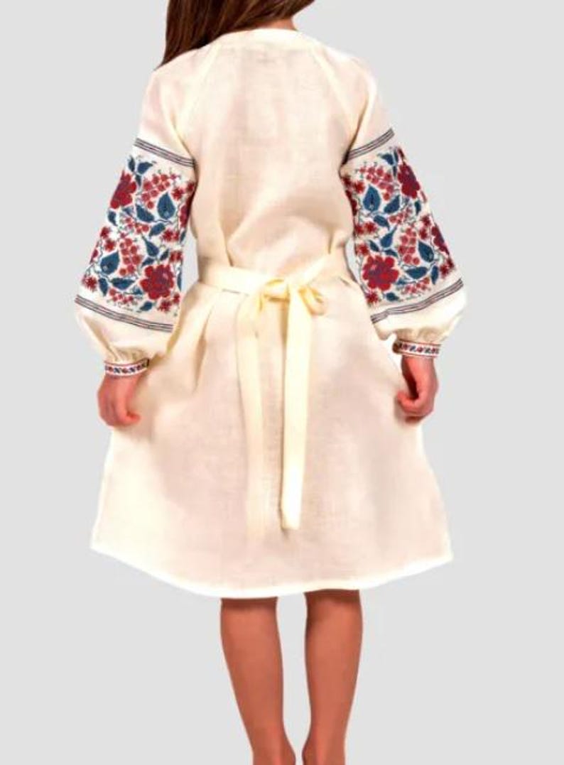 May include: A white linen dress with embroidered floral patterns in red, blue, and green on the sleeves. The dress has a tie belt at the waist.