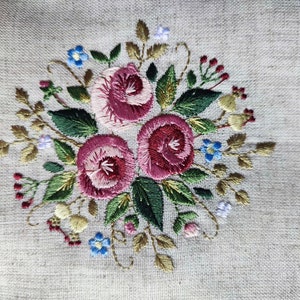 May include: Embroidered floral design featuring three large, deep pink roses surrounded by green leaves, small blue and purple flowers, and red berries. The embroidery is on a light beige linen fabric, creating a textured, handcrafted appearance.