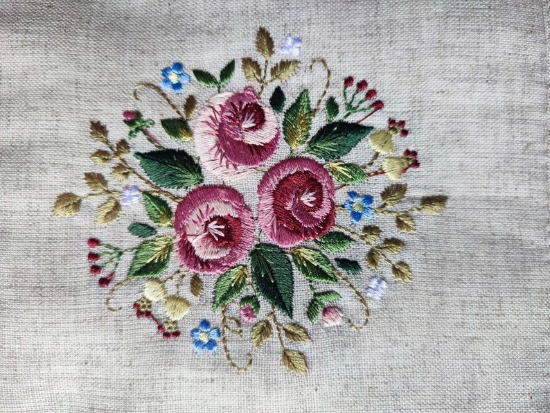 May include: Embroidered floral design featuring three large, deep pink roses surrounded by green leaves, small blue and purple flowers, and red berries. The embroidery is on a light beige linen fabric, creating a textured, handcrafted appearance.