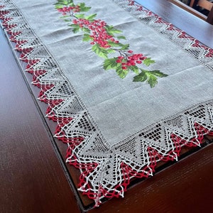 May include: A white linen table runner with red and green embroidered floral design. The runner has a delicate lace border with red accents.