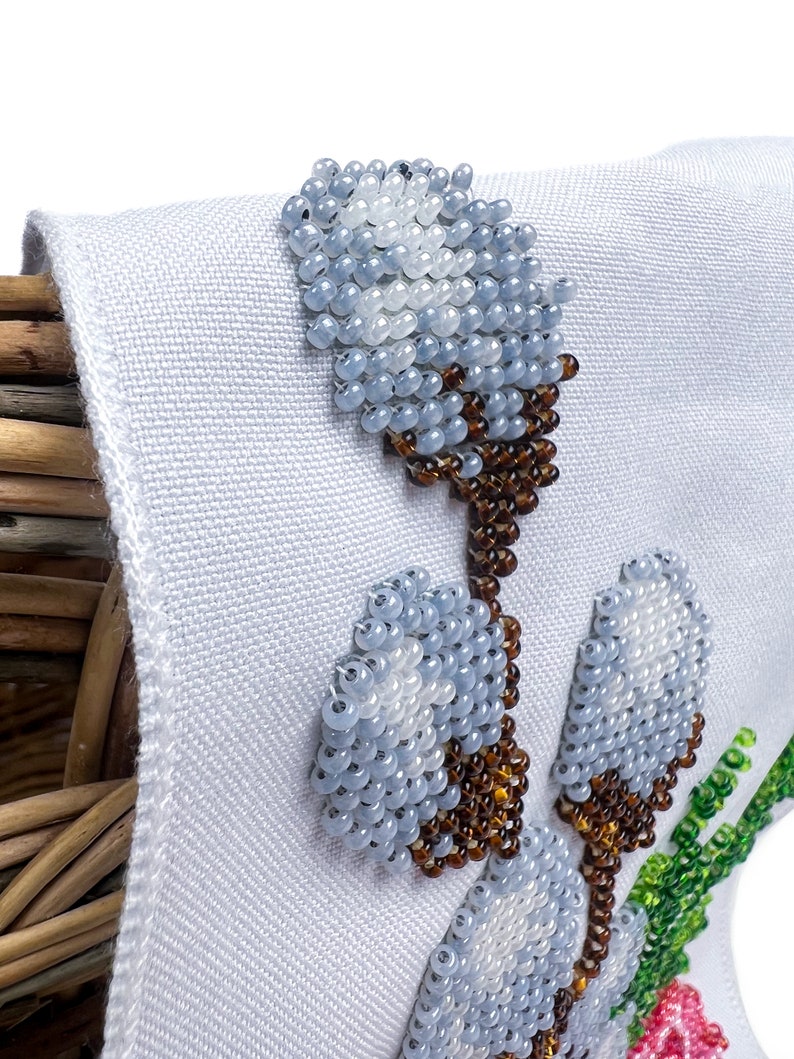May include: White fabric with a beaded design of white, blue, and brown flowers. The flowers are in a cluster and have a stem.