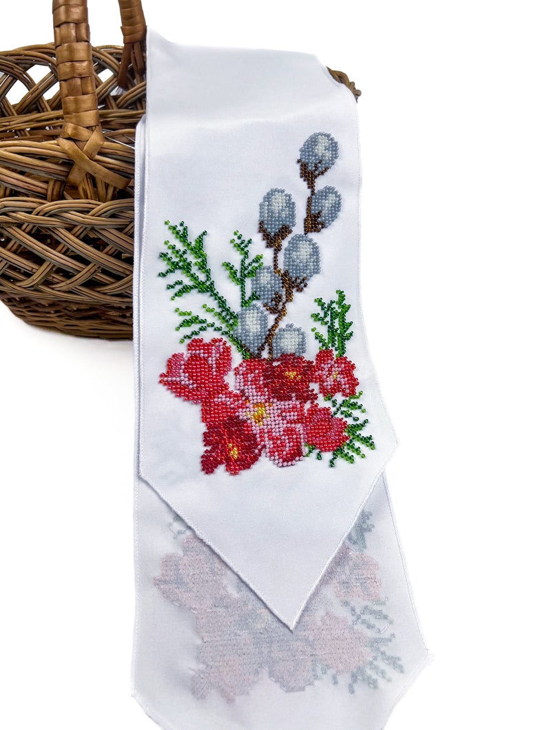May include: White fabric with a beaded embroidery design of red flowers, green leaves, and grey willow branches. The fabric is folded and draped over a wicker basket.