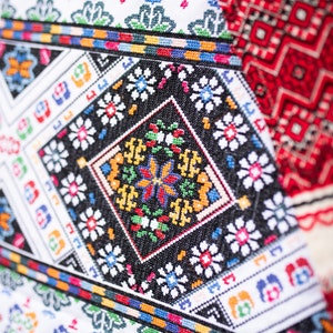 May include: Close-up of a white fabric with colorful embroidered patterns. The design features geometric shapes, flowers, and a central diamond-shaped motif with a butterfly design.
