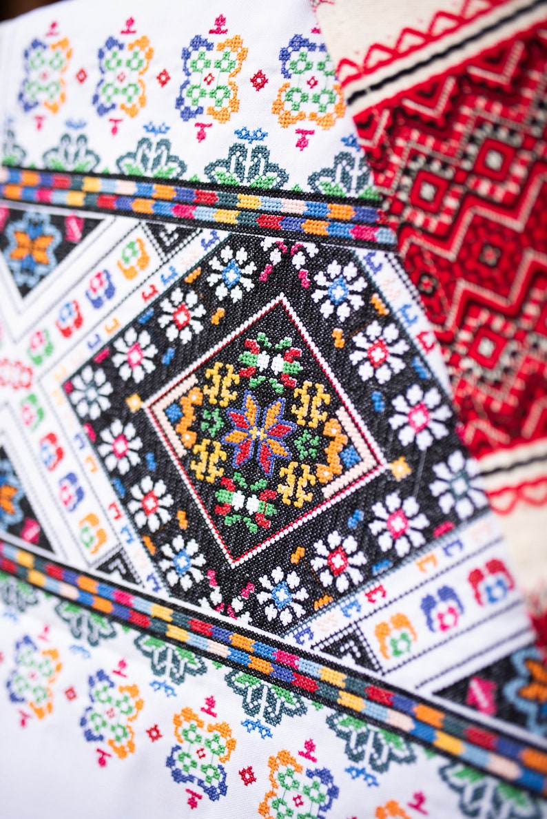 May include: Close-up of a white fabric with colorful embroidered patterns. The design features geometric shapes, flowers, and a central diamond-shaped motif with a butterfly design.