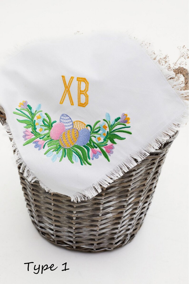 Op de afbeelding: Witte geborduurde keukendoek met een gefranst randje. De borduurwerk toont een bloemmotief met kleurrijke paaseieren en de letters "XB".