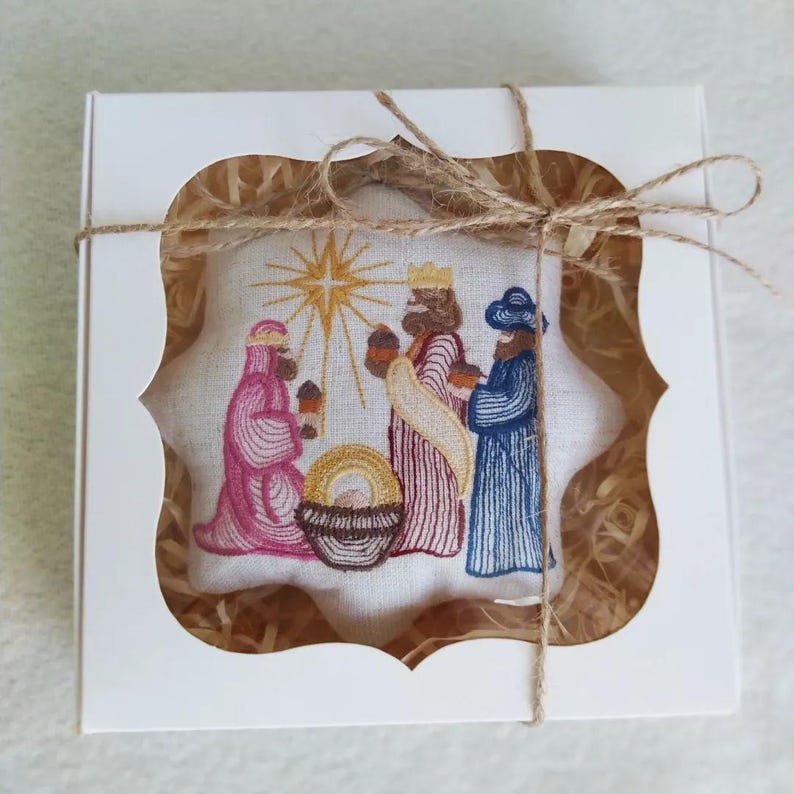 May include: Embroidered Christmas ornament featuring the three wise men, a star, and a manger scene. The ornament is displayed in a white gift box with a decorative cutout and tied with twine. The embroidery uses pink, red, blue, and gold thread.