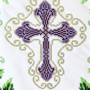 Puede incluir: Primer plano de una tela blanca con un dise&ntilde;o de punto de cruz detallado. La cruz es morada con una textura de cuentas, rodeada de filigranas doradas. Acentos florales verdes y morados en las esquinas.