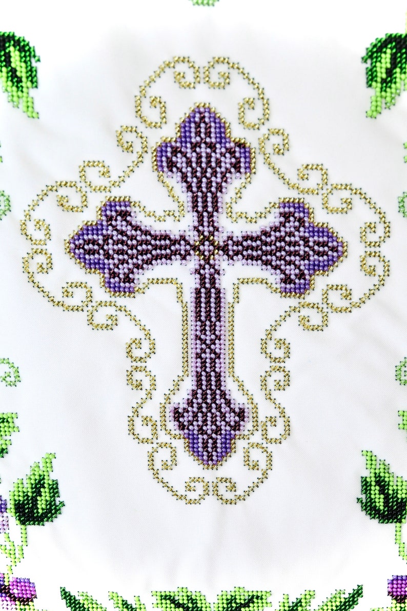 Puede incluir: Primer plano de una tela blanca con un dise&ntilde;o de punto de cruz detallado. La cruz es morada con una textura de cuentas, rodeada de filigranas doradas. Acentos florales verdes y morados en las esquinas.