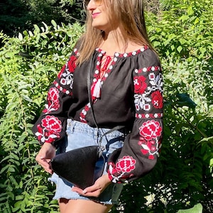 May include: A black linen blouse with red and white embroidered flowers. The blouse has a V-neckline and long sleeves. The model is wearing a black leather bag.