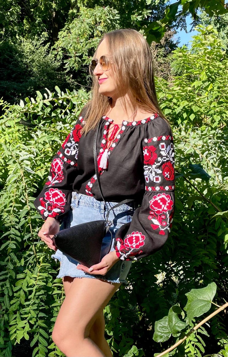 May include: A black linen blouse with red and white embroidered flowers. The blouse has a V-neckline and long sleeves. The model is wearing a black leather bag.