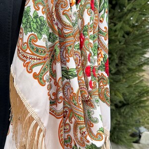 May include: A white shawl with a paisley pattern in shades of orange, green, and red. The shawl has a fringe border and is held by a person's hands.