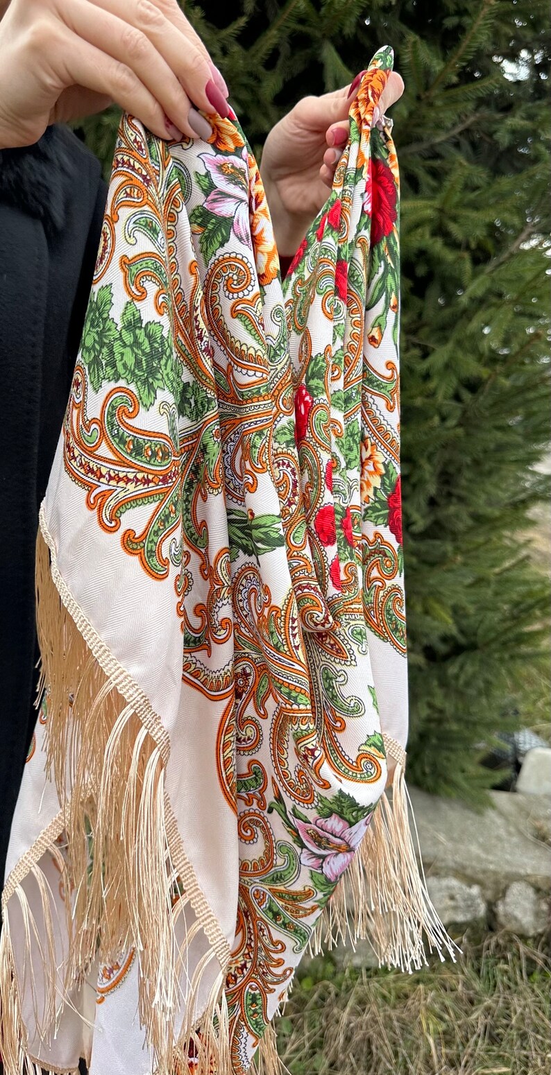 May include: A white shawl with a paisley pattern in shades of orange, green, and red. The shawl has a fringe border and is held by a person's hands.
