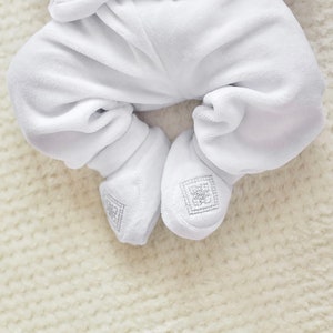 K&ouml;nnte beinhalten: Nahaufnahme eines wei&szlig;en Baby-Stramplers mit F&uuml;&szlig;en und passenden Booties aus Velours. Die Booties haben ein quadratisches, gesticktes Design. Der weiche, pl&uuml;schige Stoff suggeriert Komfort und W&auml;rme f&uuml;r ein Neugeborenes.