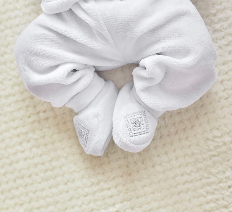 K&ouml;nnte beinhalten: Nahaufnahme eines wei&szlig;en Baby-Stramplers mit F&uuml;&szlig;en und passenden Booties aus Velours. Die Booties haben ein quadratisches, gesticktes Design. Der weiche, pl&uuml;schige Stoff suggeriert Komfort und W&auml;rme f&uuml;r ein Neugeborenes.