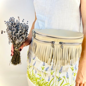 Puede incluir: Ri&ntilde;onera de cuero color crema con flecos y cremalleras plateadas. La bolsa se lleva sobre un vestido blanco con bordados de lavanda. Un ramo de lavanda seca se sostiene en la mano.