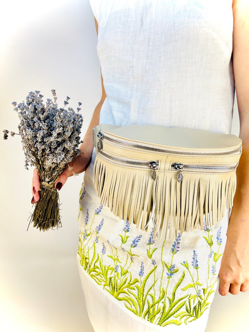 Puede incluir: Ri&ntilde;onera de cuero color crema con flecos y cremalleras plateadas. La bolsa se lleva sobre un vestido blanco con bordados de lavanda. Un ramo de lavanda seca se sostiene en la mano.
