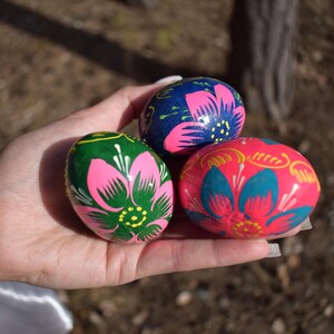 May include: Three hand-painted Easter eggs in a hand. The eggs are decorated with floral designs in pink, green, blue, and yellow. The background is blurred, with a tree trunk visible.