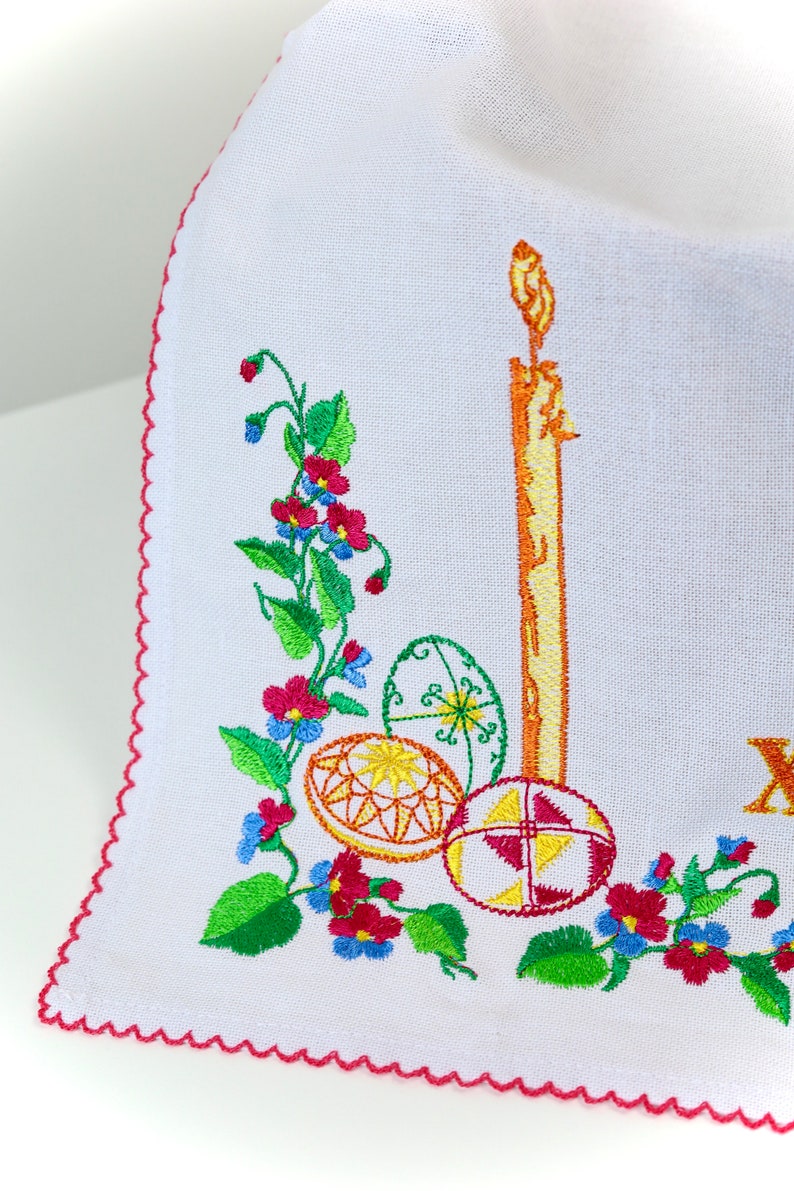 Pode incluir: Tecido branco bordado com uma borda festonada vermelha. O design apresenta uma vela, tr&ecirc;s ovos de P&aacute;scoa e uma videira com folhas verdes e flores rosa e azuis.