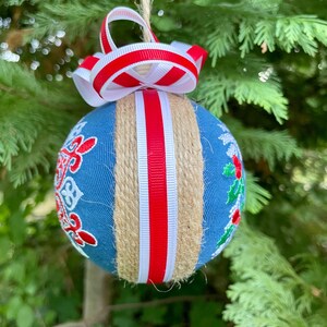K&ouml;nnte beinhalten: Ein blauer Weihnachtsbaumschmuck mit wei&szlig;em und rotem Band und Schnur. Der Schmuck ist mit einem wei&szlig;en Schneeflockenmuster und einem roten Stechpalmenzweig verziert.