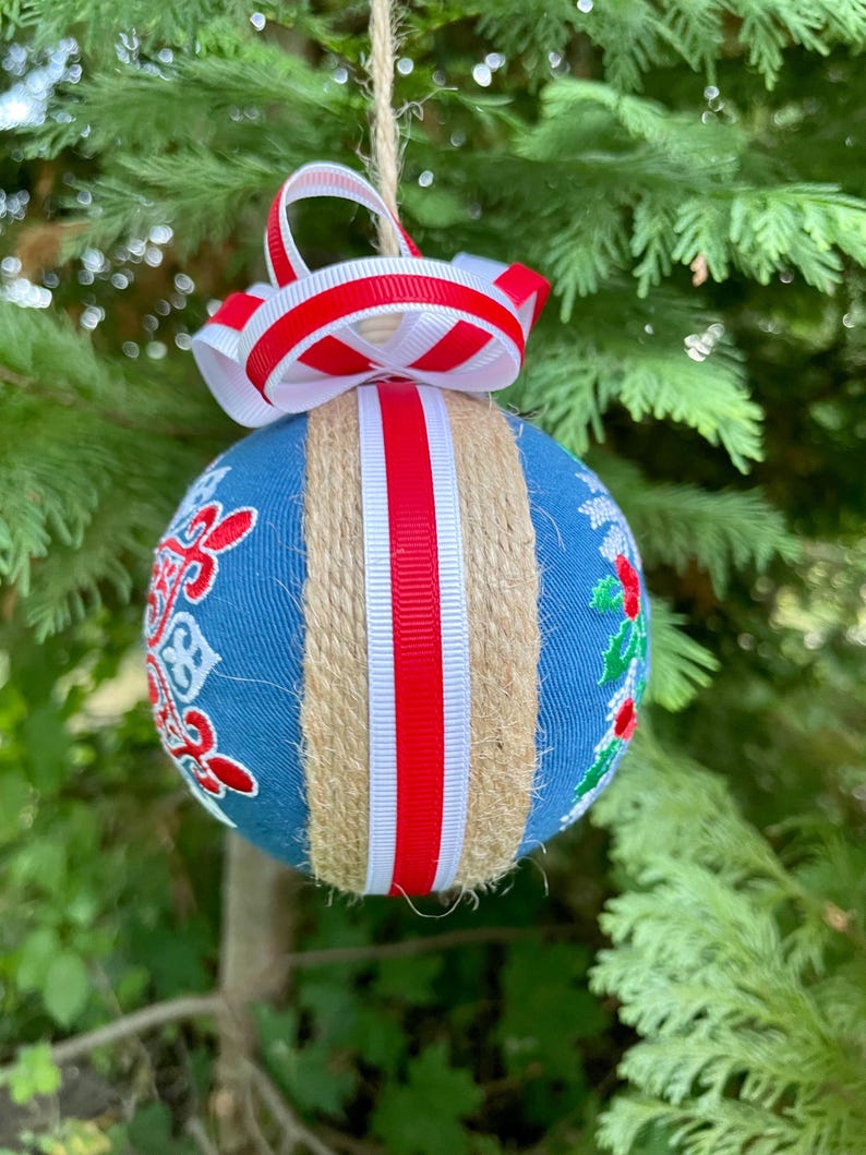 K&ouml;nnte beinhalten: Ein blauer Weihnachtsbaumschmuck mit wei&szlig;em und rotem Band und Schnur. Der Schmuck ist mit einem wei&szlig;en Schneeflockenmuster und einem roten Stechpalmenzweig verziert.