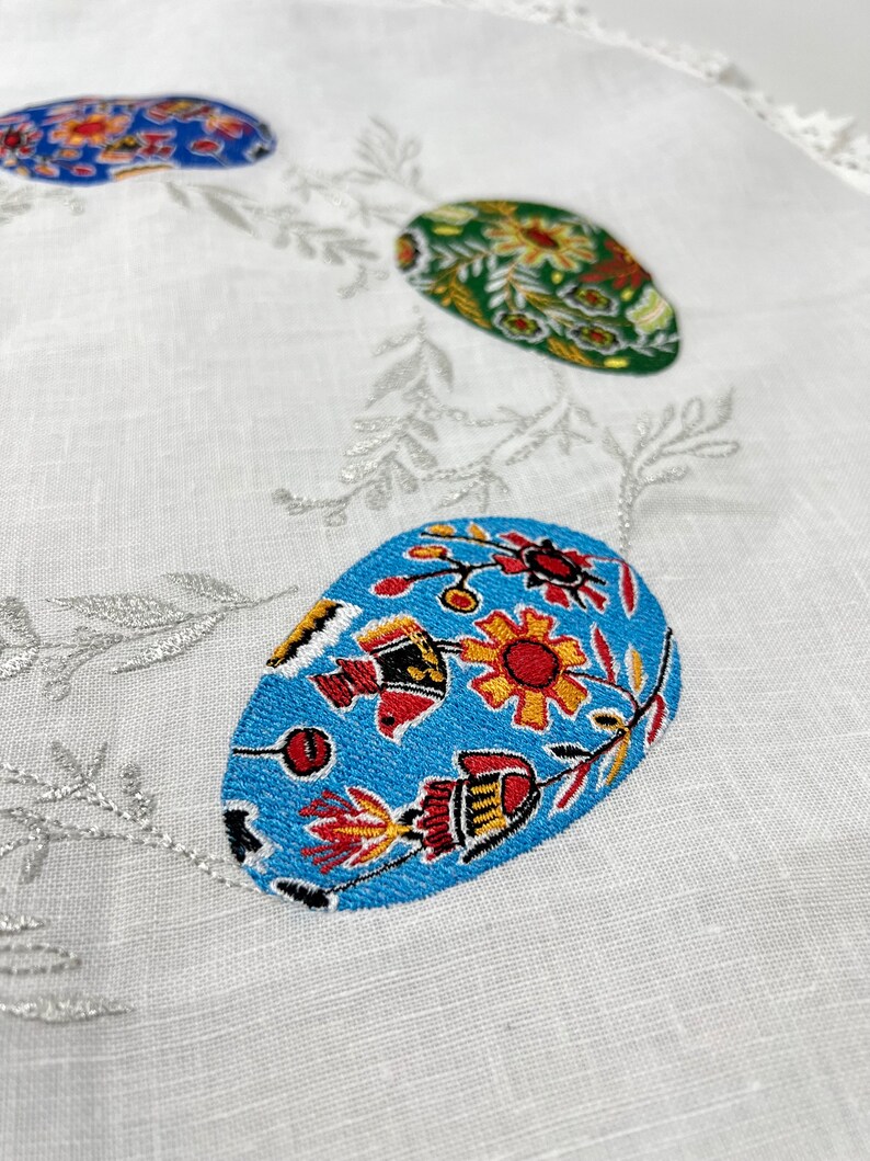 以下が含まれることがあります： 銀色のつるが刺繍された白いリネン製のテーブルクロスと、カラフルなイースターエッグ3つ。卵は青、緑、赤、黄、オレンジの色合いの花柄で飾られています。