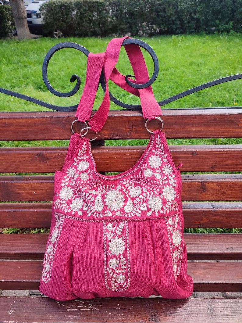 Peut inclure: Un sac cabas en tissu rose avec des motifs floraux brod&eacute;s blancs. Le sac a deux anses et un fond pliss&eacute;.