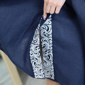 May include: A navy blue linen dress with white embroidered details along the side seams. The dress has a slit up the front and a loose, flowing silhouette.
