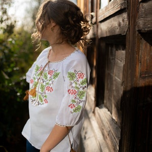 May include: A white embroidered blouse with a floral pattern and brown tassels. The blouse is worn by a person standing next to a wooden wall.