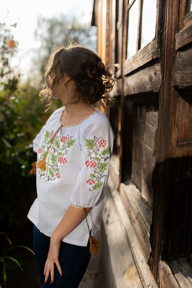 May include: A white embroidered blouse with a floral pattern and brown tassels. The blouse is worn by a person standing next to a wooden wall.