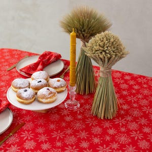 May include: A festive table setting with a red tablecloth featuring a snowflake pattern. A white plate holds several powdered sugar-dusted donuts with fruit fillings. A yellow candle sits in a crystal holder, and wheat bundles add a rustic touch.