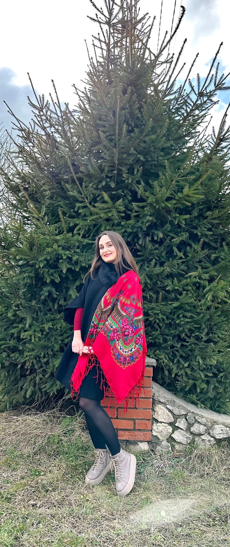 May include: A woman wearing a black coat with a fur collar and a red shawl with floral embroidery. She is standing in front of a green pine tree and wearing grey platform boots.