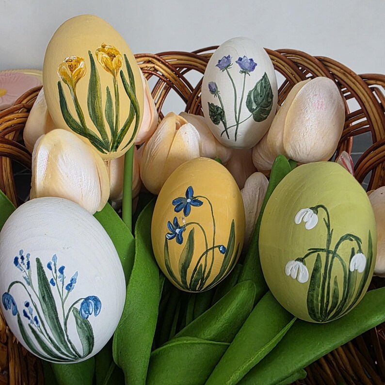May include: Hand-painted Easter eggs in a wicker basket. The eggs are decorated with floral designs in yellow, white, and green. The arrangement includes faux tulips and green leaves, creating a spring theme.