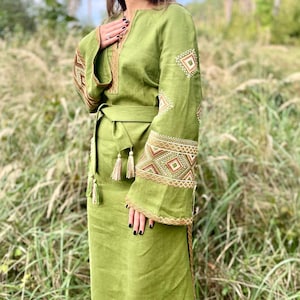 Pode incluir: Um vestido longo verde-oliva com mangas compridas e cinto a condizer. As mangas e o decote s&atilde;o adornados com bordados intrincados em tons de castanho e dourado. O vestido tem uma saia fluida e &eacute; feito de um tecido leve.