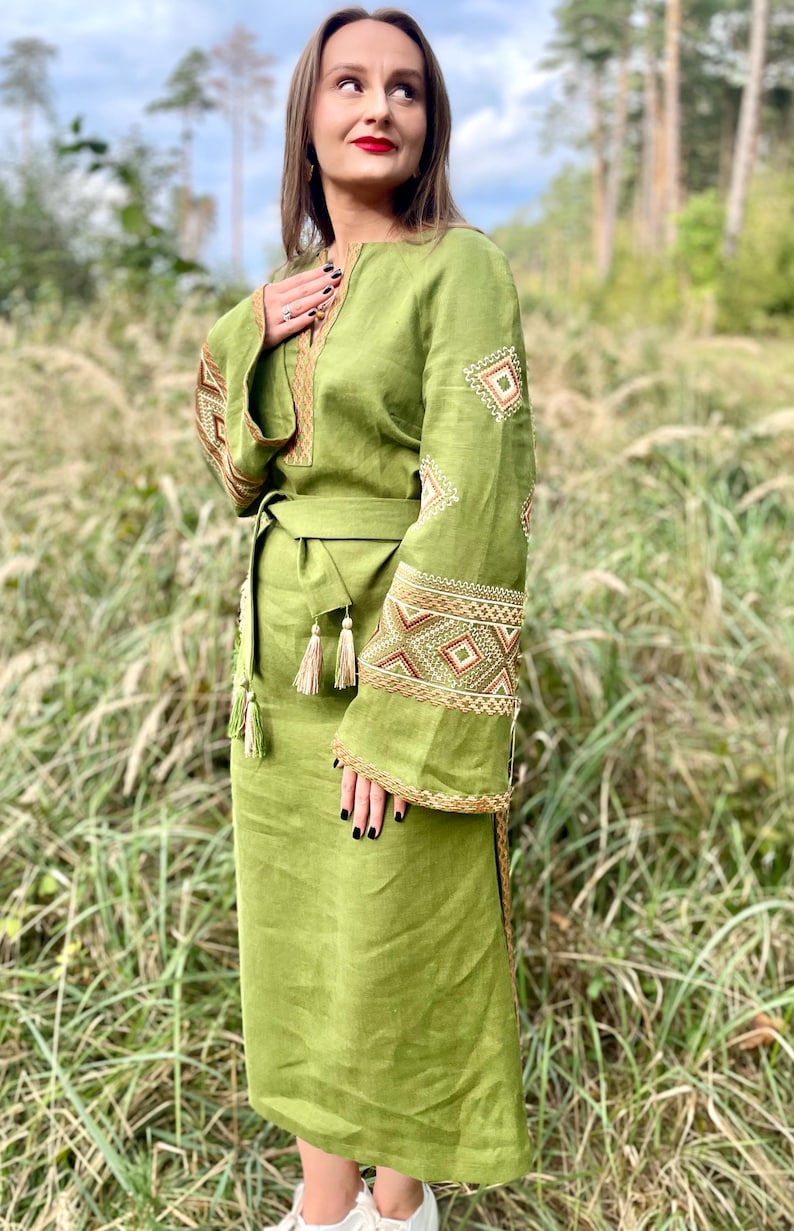 Pode incluir: Um vestido longo verde-oliva com mangas compridas e cinto a condizer. As mangas e o decote s&atilde;o adornados com bordados intrincados em tons de castanho e dourado. O vestido tem uma saia fluida e &eacute; feito de um tecido leve.
