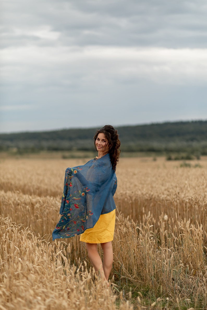 May include: A woman in a yellow dress and a blue floral scarf stands in a field of wheat. She is looking over her shoulder and smiling.