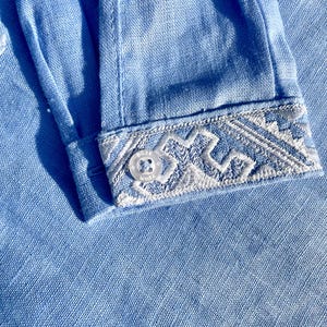 May include: Close-up of a light blue linen shirt sleeve. The cuff features intricate white embroidery with a geometric design. A small, clear button is visible on the cuff. The fabric has a textured appearance.