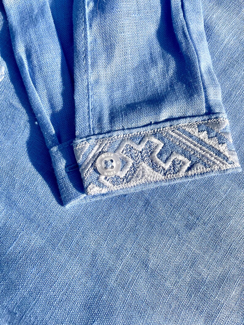 May include: Close-up of a light blue linen shirt sleeve. The cuff features intricate white embroidery with a geometric design. A small, clear button is visible on the cuff. The fabric has a textured appearance.